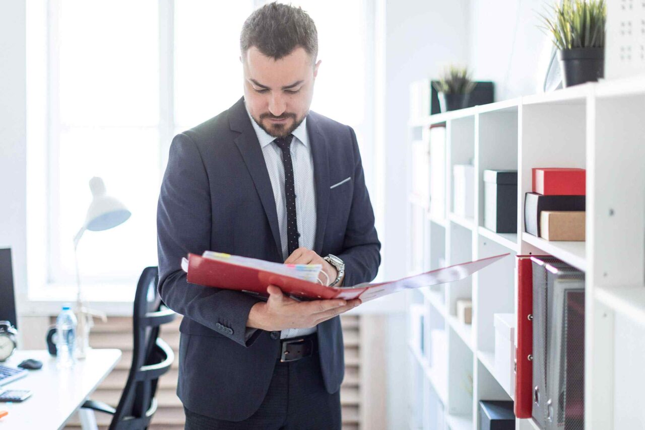 Businessman in a suit holding a red folder in a bright office setting.